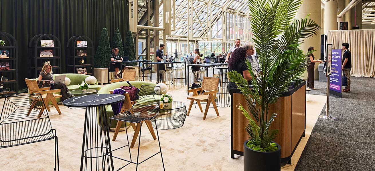 Attendees seated at tables in a bright conference venue with large windows and an open gathering area.A lounge area with tables, chairs and plants at a conference