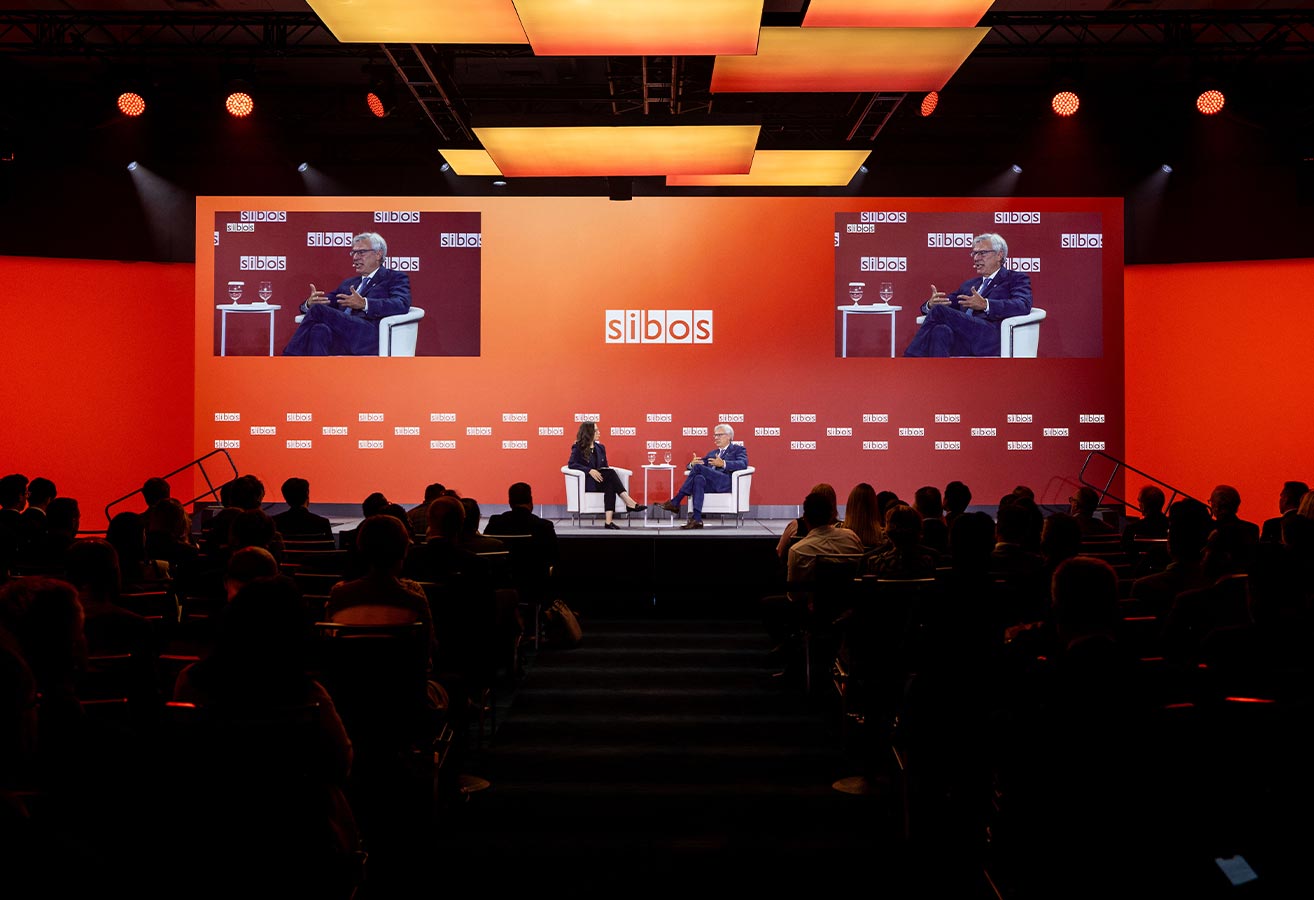 Large audience seated in a conference hall facing an illuminated stage.