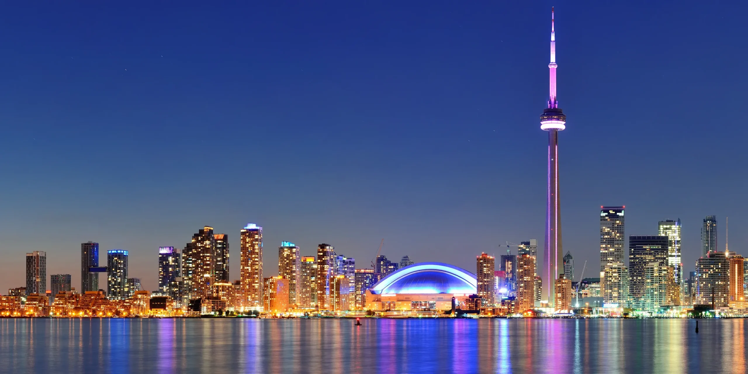 The Toronto skyline at night, with the CN Tower and Rogers Centre lit up and reflected in Lake Ontario.