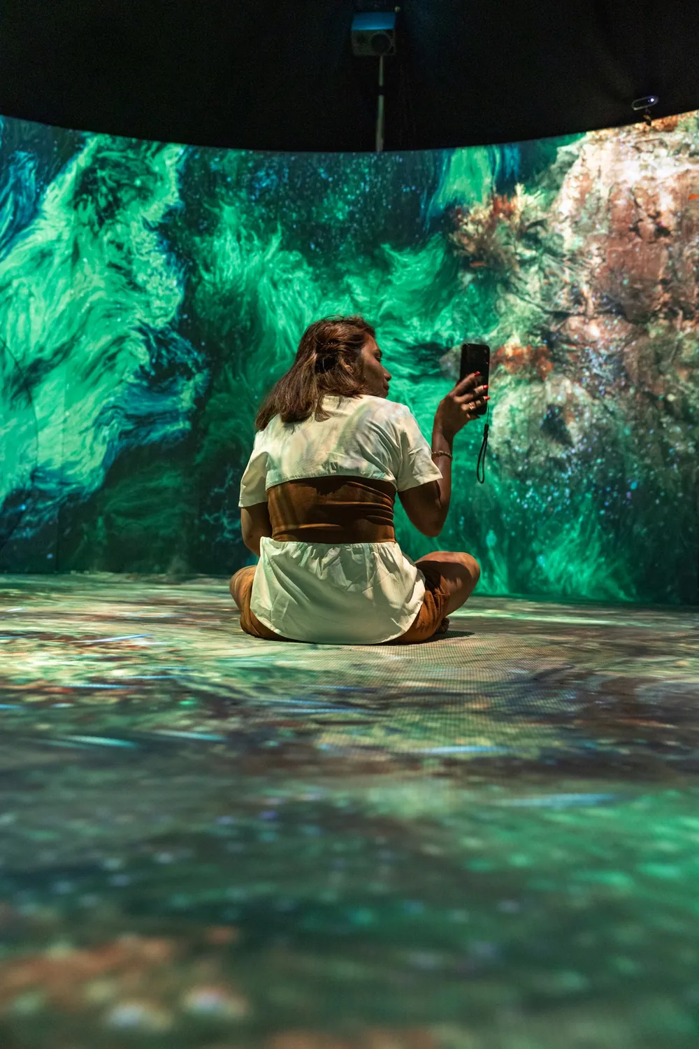 A visitor sits on the floor of an immersive digital art installation, photographing the glowing green projections