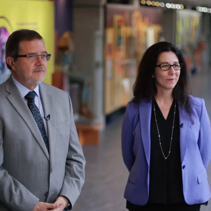 Dr. Maurice Bitran and Catherine Paisley standing together in an indoor gallery, wearing a grey suit and purple blazer.
