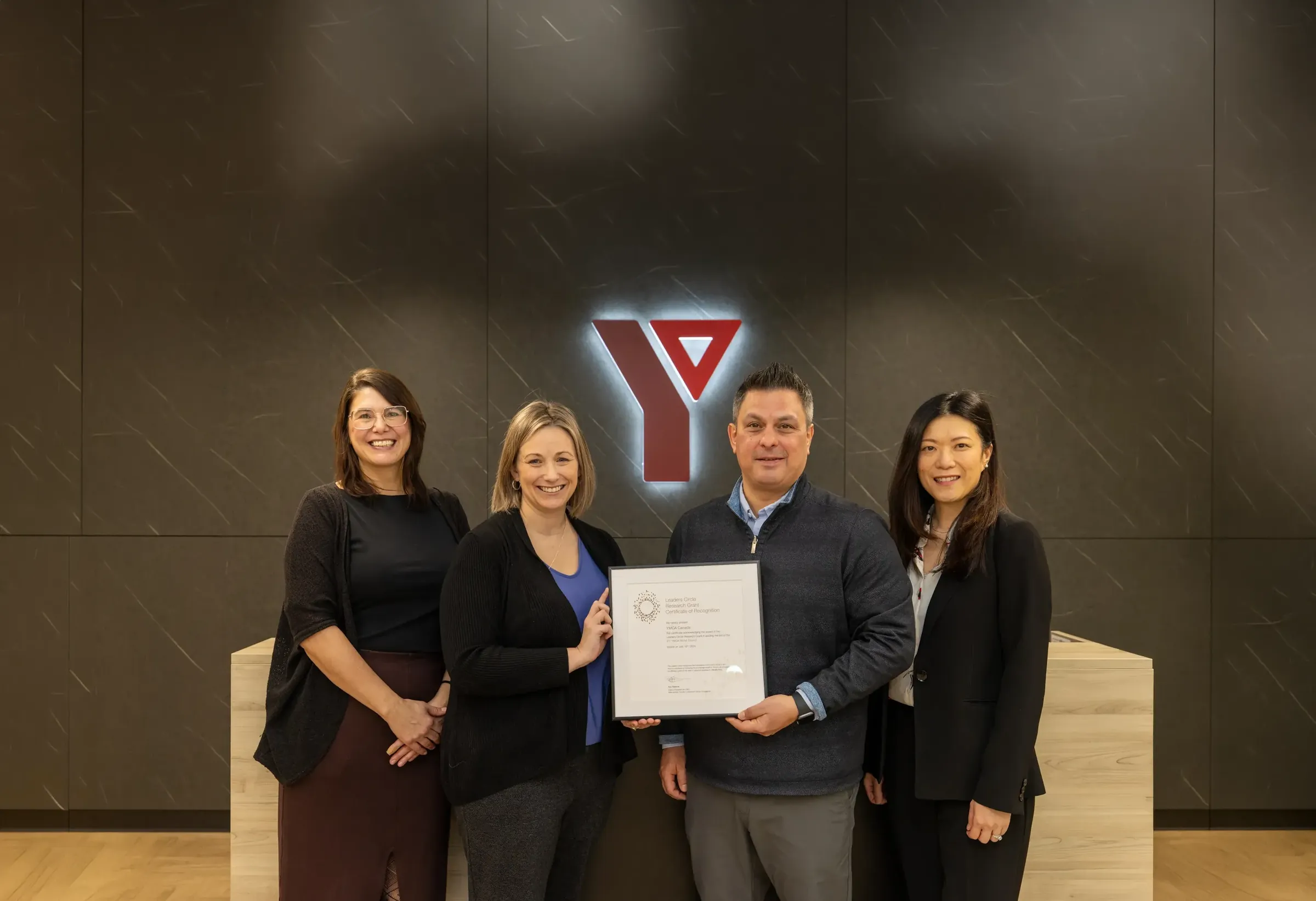 Four people stand in front of a lit YMCA logo, one holding a Leaders Circle Research Grant certificate of recognition for YMCA Canada