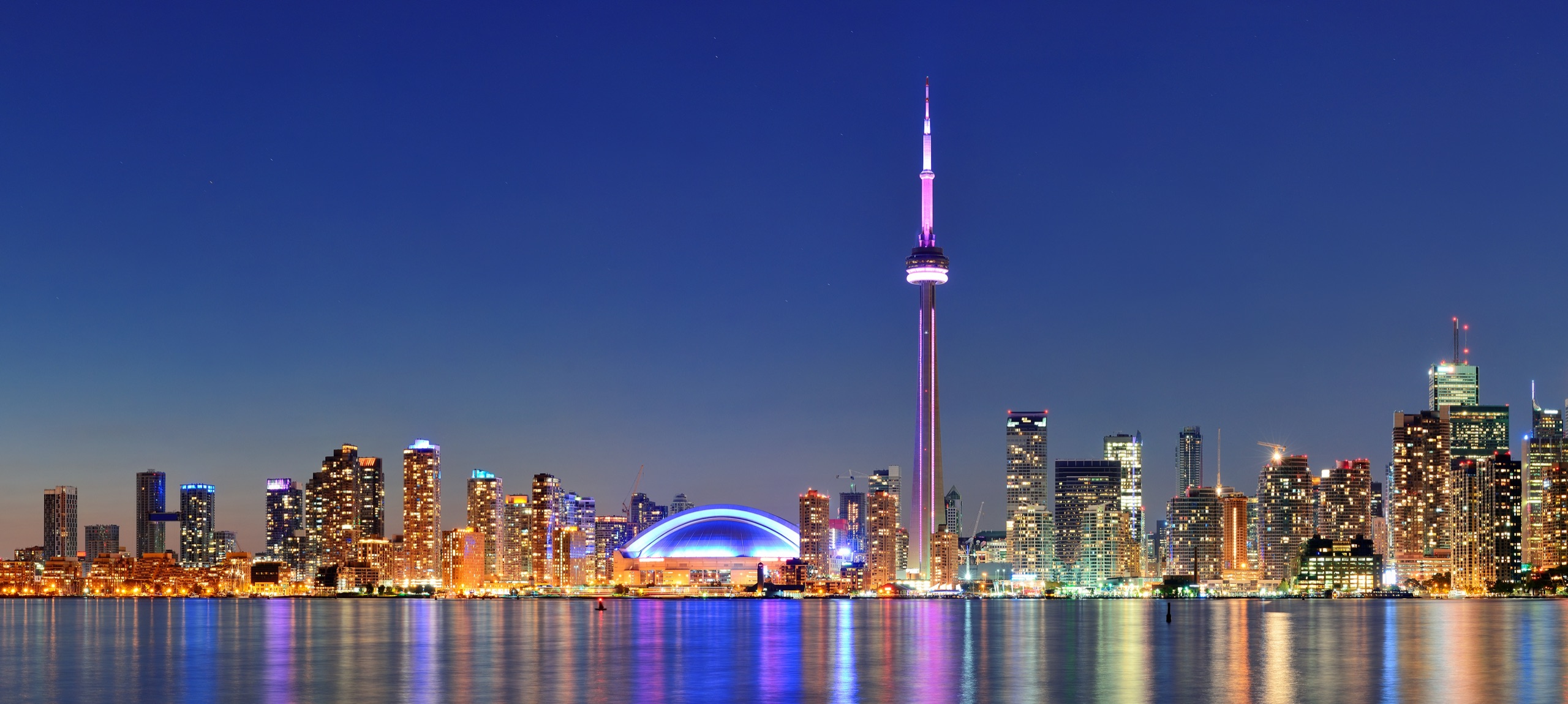 Toronto skyline illuminated at night with the CN Tower rising above waterfront buildings.